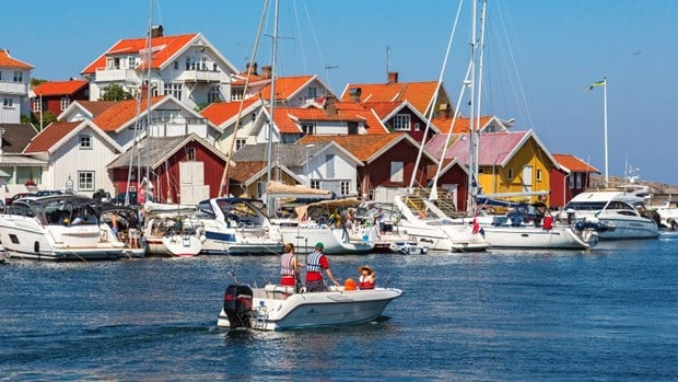 En familj är ute i båten vid en hamn bland många båtar, fina hus och svenska flaggor